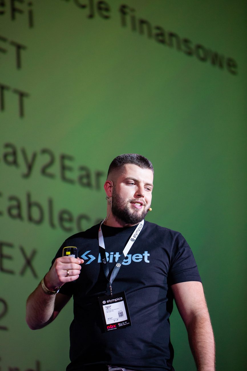 Igor Haleta on the NBX Warsaw 2024 main stage with a headset microphone, wearing a Bitget speaker shirt